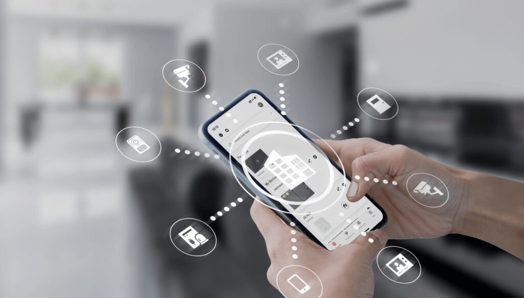 Person holding a smartphone with digital smart home icons floating around it in a blurred modern interior.